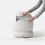 A person wearing a gray long-sleeve top is opening the lid of a white, rounded countertop trash bin or compactor on a clean white background.