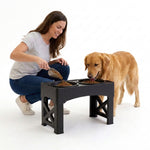 A woman crouches beside a golden retriever dog using a black elevated double pet feeder. The dog is eating from one bowl while the woman pours kibble into the second bowl on a clean white background.