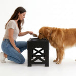 A woman kneels on a white studio floor feeding a golden retriever dog from a black elevated dog bowl. The dog is drinking from the bowl while the woman holds a food scoop nearby.