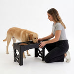 A woman kneels beside a golden retriever dog using a black elevated dog feeder with two bowls, photographed on a clean white studio background.